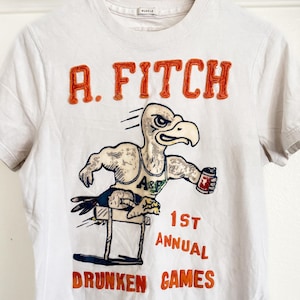 May include: White t-shirt with the text "R. Fitch" in orange, above a cartoon eagle holding a can and jumping a hurdle. The text "1st Annual Drunken Games" is below the eagle in orange.