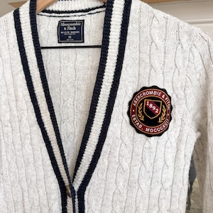 Op de afbeelding: Crèmekleurig kabelgebreid vest met marineblauwe en witte gestreepte bies. Voorzien van een patch met de tekst "Abercrombie & Fitch" en het jaartal "1892". Het vest hangt aan een houten kleerhanger.