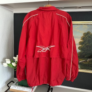 Puede incluir: Chaqueta cortavientos Reebok roja con ribete blanco y el logotipo de Reebok en la espalda. La chaqueta está colgada de una percha de madera. La chaqueta tiene un cordón en la parte inferior.