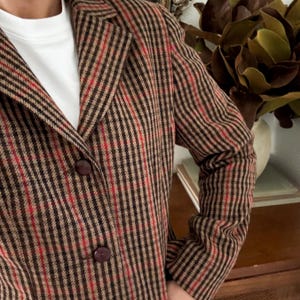 May include: A blazer featuring a brown, tan, and red vertical striped pattern. The garment has a notched lapel, long sleeves, and two dark brown circular buttons centered on the front. A plain white crew neck shirt is visible underneath the blazer.