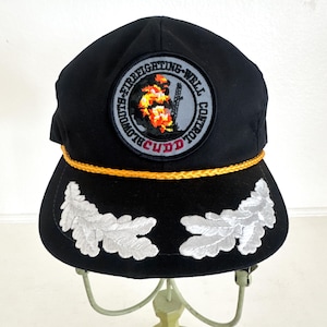 May include: Black baseball cap featuring a patch with the text "Blowouts-Firefighting-Well Control" and a fire graphic. The cap has gold rope detailing and white leaf embroidery.