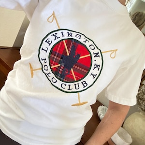 以下が含まれることがあります： 背面にレキシントンポロクラブのエンブレムが付いた白いTシャツ。エンブレムには、ネイビーブルーのシルエット、赤と黒のチェック柄、そして「LEXINGTON N.Y. POLO CLUB」の文字が金色のアクセントで円形にデザインされています。