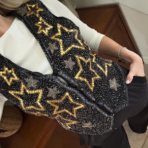 May include: Black vest embellished with gold and silver star-shaped sequins. The vest has a dark background and a V-neck design. The stars are outlined in gold sequins and filled with smaller silver sequins.