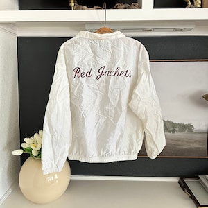 May include: A white windbreaker jacket with the words "Red Jackets" embroidered in burgundy script across the back. The jacket has a collared neckline and a drawstring hem. The jacket is hanging on a wooden hanger.
