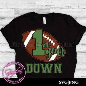 May include: Black t-shirt with a green number one and the words "1st year down" on a brown and white football graphic.