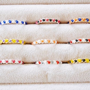 May include: Nine colorful beaded rings displayed in a velvet ring box. The rings are made with small beads in various colors, including yellow, orange, red, blue, black, and white.
