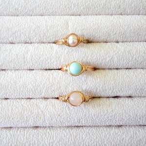 May include: Three gold wire wrapped rings with different stones. The top ring has a white pearl, the middle ring has a light blue stone, and the bottom ring has a light pink stone.