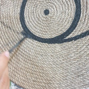 May include: A close-up of a round, woven jute rug with a black painted design of a circle and a dot in the center.