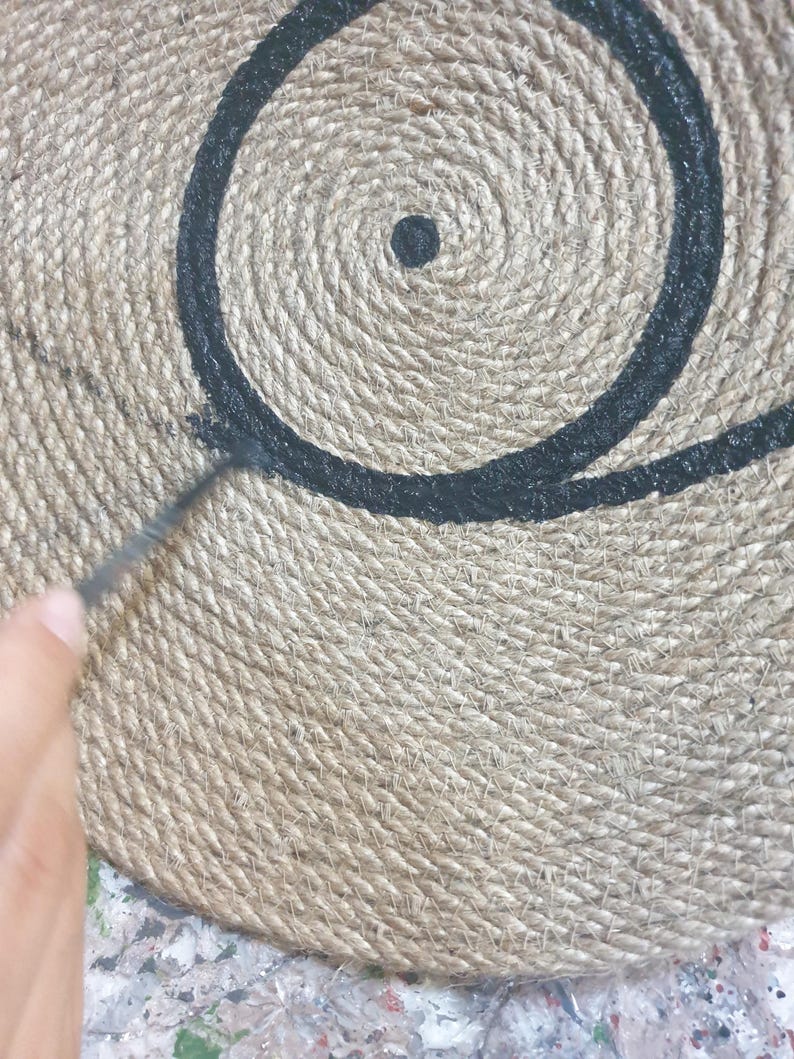 May include: A close-up of a round, woven jute rug with a black painted design of a circle and a dot in the center.