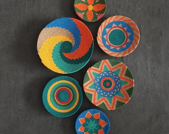 May include: Six colourful woven wall baskets with geometric designs in shades of blue, green, orange, and brown.