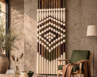 Op de afbeelding: Een verticale wandhanger met een reeks lange, dunne staven omwikkeld met crèmekleurig, bruin en olijfgroen garen. Het ontwerp bevat rijen crèmekleurige bolvormige kralen en eindigt met lange, rechte crèmekleurige franjes aan de onderzijde.
