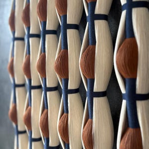 May include: Close-up of a decorative wall hanging. The design features vertical rows of cream-coloured, elongated shapes with a dark brown, rounded centre. Dark blue bands encircle the shapes, creating a repeating pattern. The background is a muted grey.