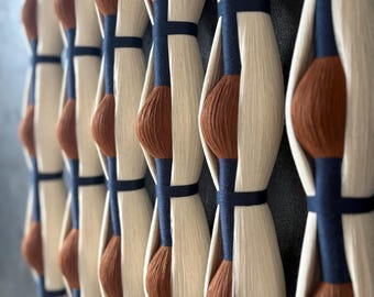 May include: Close-up of a decorative wall hanging. The design features vertical rows of cream-coloured, elongated shapes with a dark brown, rounded centre. Dark blue bands encircle the shapes, creating a repeating pattern. The background is a muted grey.