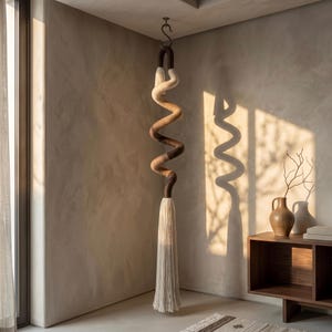 May include: A hanging spiral sculpture made of wrapped yarn in shades of cream, tan, and dark brown, suspended from a metal ceiling hook. The sculpture features a long, thick fringe tassel at the bottom, transitioning from dark brown to cream.