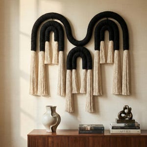 May include: A large, modern wall hanging featuring a black and cream color scheme. The design incorporates arched shapes with long, cream-colored tassels. The piece is mounted on a neutral-toned wall, creating a striking visual contrast.
