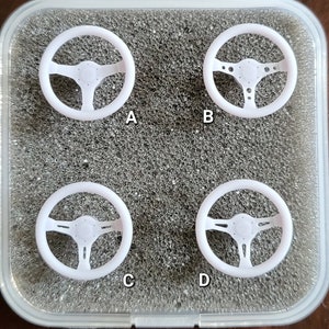 May include: Four white steering wheels, labeled A, B, C, and D, are displayed in a clear plastic container with a gray foam interior.