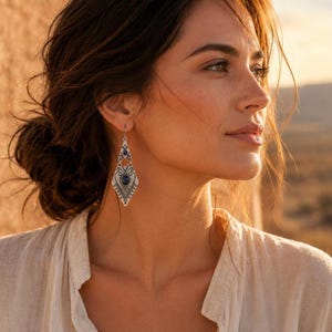 May include: A silver-toned dangle earring featuring a diamond-shaped metal frame with intricate black etched patterns and two circular dark blue stones set vertically within the center of the geometric design, suspended from a simple metal hook.
