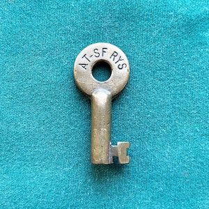 May include: A vintage brass key with a circular head and a rectangular bit. The head of the key has the text "AT-SF RYS" in a circular arrangement. The key is set against a teal fabric background.
