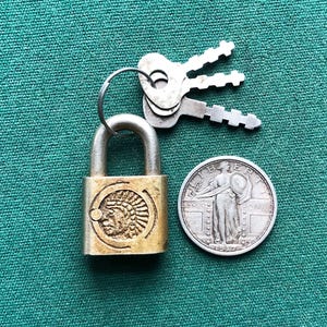 May include: A brass padlock with a keyhole and a design of a Native American head on the front. The padlock is attached to three keys and is next to a silver 1917 quarter.