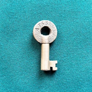 May include: A metallic skeleton key with a circular bow featuring the embossed text A&S RY. The key has a cylindrical shank and a notched bit at the bottom. The surface of the bow has a textured, aged appearance while the shank is smooth and light gray.