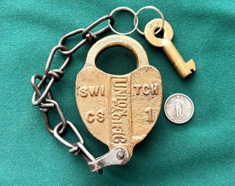 May include: A brass heart-shaped Union Pacific switch padlock with a metal chain and a brass key attached to a ring. The lock features the embossed text UNION PACIFIC SWITCH CS 1. A silver-colored coin is positioned next to the padlock for scale.
