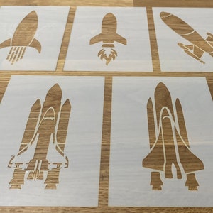 Peut inclure: Cinq pochoirs blancs de différentes fusées, y compris une navette spatiale, sur une surface en bois.
