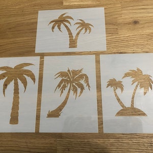May include: Four white stencils of palm trees, each with a different design. The stencils are on a wooden surface.
