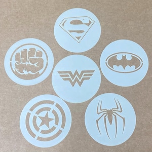 Op de afbeelding: Een set van zes ronde witte sjablonen met diverse superhelden-emblemen, waaronder een vuist, een ruitvormige S, een vleermuissilhouet, een dubbel W-logo, een ster binnen concentrische cirkels en een spingrafiek, gerangschikt in een groep.