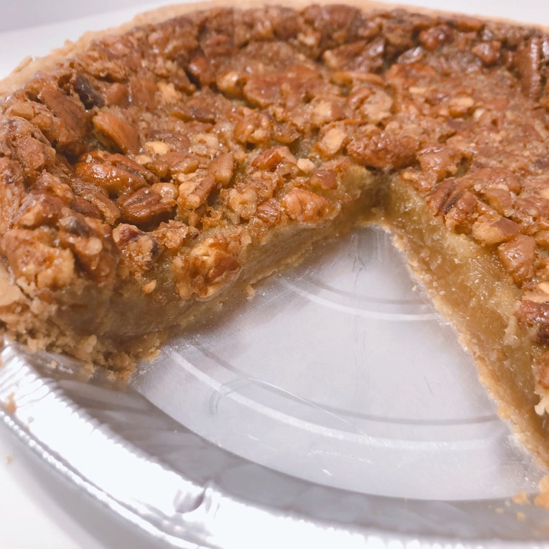 K&ouml;nnte beinhalten: Ein Pecan Pie mit einer goldbraunen Kruste und einem fehlenden St&uuml;ck, das die s&uuml;&szlig;e F&uuml;llung freilegt. Der Kuchen befindet sich in einer silbernen Alu-Tortenform.
