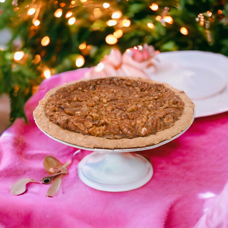 K&ouml;nnte beinhalten: Ein Pecan Pie auf einem wei&szlig;en Kuchenst&auml;nder mit einer rosa Tischdecke darunter. Der Kuchen steht auf einem wei&szlig;en Kuchenst&auml;nder mit einer rosa Tischdecke darunter. Der Kuchen ist von einem verschwommenen Hintergrund eines Weihnachtsbaums mit Lichtern umgeben.