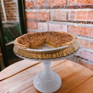 K&ouml;nnte beinhalten: Ein Pecan Pie mit einem fehlenden St&uuml;ck, pr&auml;sentiert auf einem wei&szlig;en Kuchenst&auml;nder mit einer goldenen Papierunterlage. Der Kuchen befindet sich in einer silbernen Alu-Tortenform.