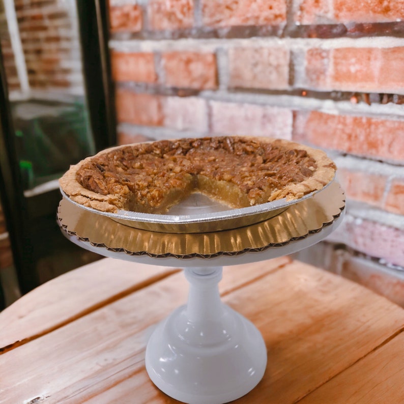 K&ouml;nnte beinhalten: Ein Pecan Pie mit einem fehlenden St&uuml;ck, pr&auml;sentiert auf einem wei&szlig;en Kuchenst&auml;nder mit einer goldenen Papierunterlage. Der Kuchen befindet sich in einer silbernen Alu-Tortenform.