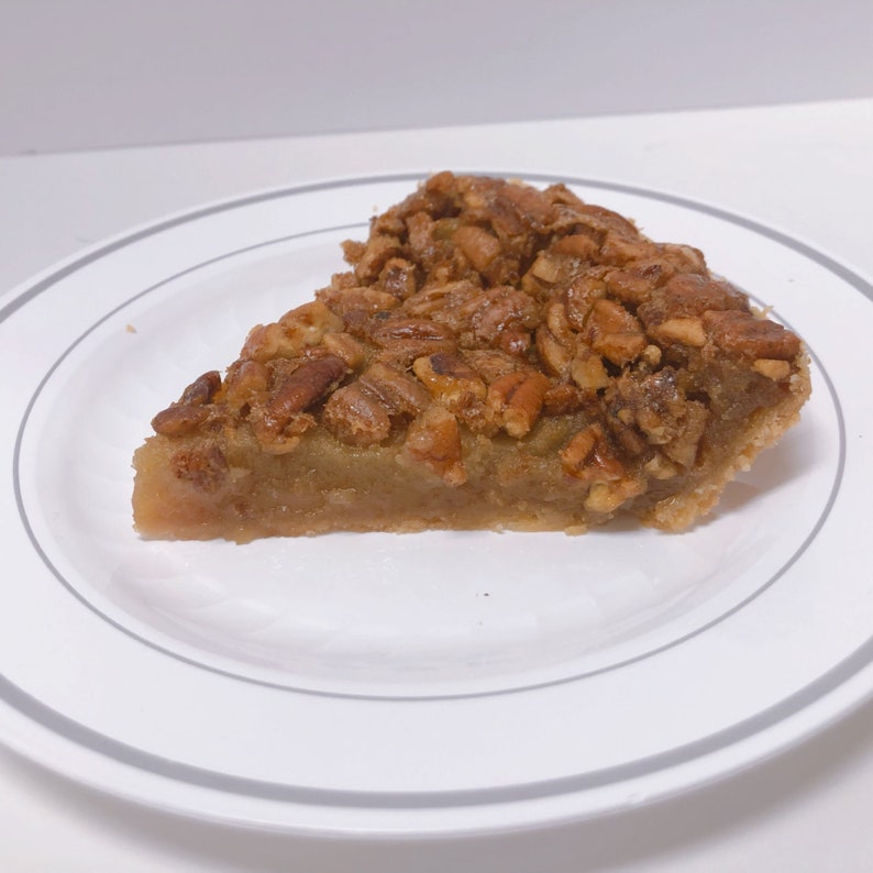 K&ouml;nnte beinhalten: Ein St&uuml;ck Pecan Pie auf einem wei&szlig;en Teller mit grauem Rand. Der Kuchen ist mit einer hellbraunen F&uuml;llung gef&uuml;llt und mit einer gro&szlig;z&uuml;gigen Menge Pecan&uuml;sse belegt.