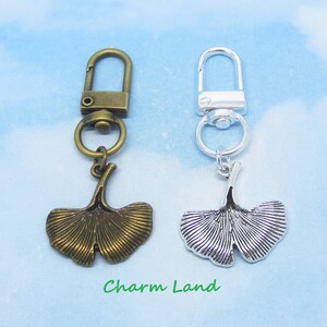 May include: Two metal keychains with ginkgo leaf charms. One keychain has a bronze finish, the other a silver finish. The ginkgo leaf charms are detailed with veining. The text "Charm Land" is printed in green.