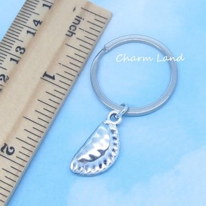 May include: Silver-colored keychain featuring a dumpling charm. The charm is detailed with a crimped edge and a curved shape. The keychain ring is attached to the charm with a small loop. The text "Charm Land" is visible on the image.