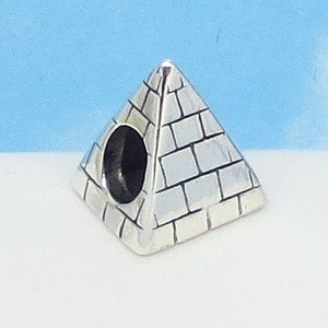 May include: A silver-colored pyramid charm with a detailed brick pattern. The charm features a large, dark hole on one side, and is set against a light blue and white background. The pyramid is a three-dimensional shape.