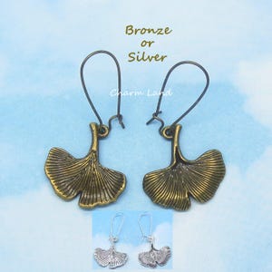 May include: A pair of bronze-colored earrings with a ginkgo leaf design. The earrings have a hook closure and are displayed with a smaller pair of silver earrings. The text "Bronze or Silver" and "Charm Land" are visible.