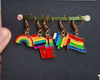 Puede incluir: Conjunto de cinco marcadores de punto de colores arcoíris con diferentes diseños, que incluyen un arcoíris, un corazón, una barra y una bandera. Los marcadores están unidos a una cinta verde y se muestran en una tarjeta negra con el texto "The Wool Cauldron" y la información de contacto.