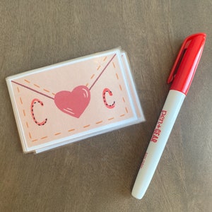 Op de afbeelding: Een stapel blanco kaarten met een roze hart en de letter "C" op elke kaart. De kaarten zijn versierd met een gestikt envelopontwerp. Een rode pen+gear-marker ligt naast de kaarten.