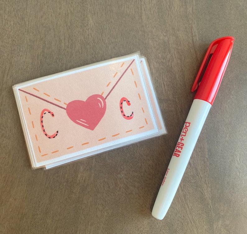 Op de afbeelding: Een stapel blanco kaarten met een roze hart en de letter "C" op elke kaart. De kaarten zijn versierd met een gestikt envelopontwerp. Een rode pen+gear-marker ligt naast de kaarten.