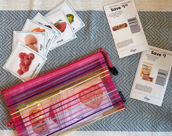 Puede incluir: Una bolsa de malla rosa con cremallera y rayas arcoíris que contiene tarjetas de memoria con imágenes y palabras para diferentes alimentos, como frambuesas, zanahorias, huevos, bistec, galletas, cereales y chocolate. Dos cupones de Kroger son visibles en el fondo, uno por 1 € y otro por 110 €.