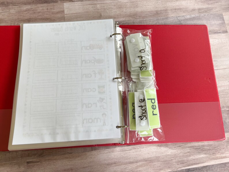 May include: A red binder with two clear plastic pockets containing green and white cards. The cards have the words "short a", "short e", and "red" written on them. The binder has a white page with the words " CVC Words" written on it.