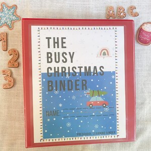 May include: Red binder with a white cover that says "The Busy Christmas Binder" with a red car carrying a Christmas tree on a blue background with snowflakes. The binder is labeled "Preschool Learning Binder".
