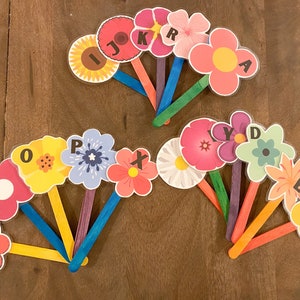 K&ouml;nnte beinhalten: Ein Satz von 14 bunten, blumenf&ouml;rmigen Karten mit darauf gedruckten Buchstaben des Alphabets. Die Karten sind an Holzst&auml;bchen befestigt.