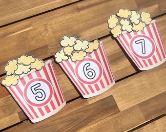 May include: Three red and white striped popcorn containers with numbers 5, 6, and 7 on them. Each container is filled with yellow popcorn.