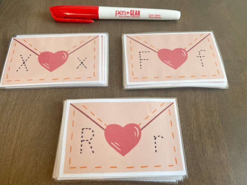 Op de afbeelding: Drie stapels kaarten met een roze envelopontwerp. Elke kaart heeft een hart en een letter in de envelop. De letters zijn X, F en R.