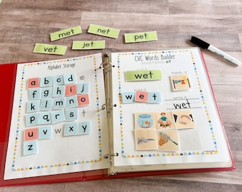 Op de afbeelding: Een rode map met een witte pagina open naar een CVC Words Builder-activiteit. De pagina toont het woord "wet" met een afbeelding van een natte hond, een viskom en een net. De linkerpagina heeft een sectie voor het opslaan van het alfabet met lettertegels.