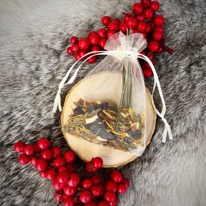 May include: A small, sheer, drawstring bag filled with dried herbs and spices, resting on a wooden slice. The bag is tied with a white ribbon. Red berries and greenery surround the bag, set against a gray, textured background.