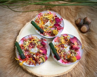 May include: Three round candles in silver containers, each adorned with dried flowers and a green stick. The candles are arranged on a wooden slice, with a backdrop of faux fur. The candles feature pink wax and a variety of colorful dried botanicals.