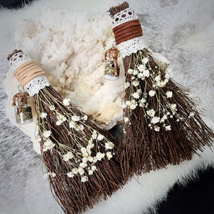 May include: Two decorative brooms made of brown twigs, adorned with white baby's breath flowers. Each broom features a lace and leather wrap at the handle, and a small glass vial charm filled with herbs. The brooms are displayed on a white fur-like surface.